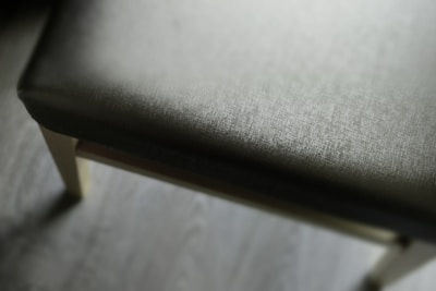 A close-up view of a padded chair seat with a texture similar to fabric. The seat is in focus while the background is blurred. The chair legs are wooden, and the floor beneath is made of light-colored wood.