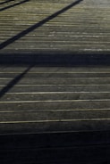 A wooden deck surface with straight horizontal lines accentuated by the planks. Shadows of an unseen structure create diagonal patterns across the deck, giving it a textured appearance.