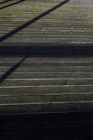 A wooden deck surface with straight horizontal lines accentuated by the planks. Shadows of an unseen structure create diagonal patterns across the deck, giving it a textured appearance.