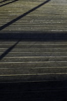 A wooden deck surface with straight horizontal lines accentuated by the planks. Shadows of an unseen structure create diagonal patterns across the deck, giving it a textured appearance.
