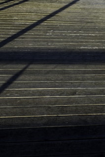A wooden deck surface with straight horizontal lines accentuated by the planks. Shadows of an unseen structure create diagonal patterns across the deck, giving it a textured appearance.