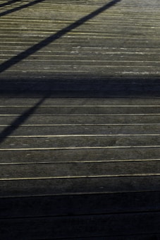 A wooden deck surface with straight horizontal lines accentuated by the planks. Shadows of an unseen structure create diagonal patterns across the deck, giving it a textured appearance.