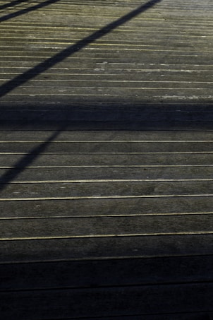 A wooden deck surface with straight horizontal lines accentuated by the planks. Shadows of an unseen structure create diagonal patterns across the deck, giving it a textured appearance.