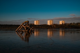 A serene landscape featuring three identical residential buildings in the distance, with a clear reflection on the water in the foreground. The scene is enhanced by a prominent driftwood structure emerging from the ice-covered lake. Trees and bushes line the horizon under a deep blue, cloud-streaked sky.