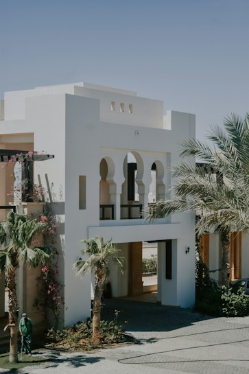 A modern building with a minimalist design featuring white walls and large arches. Palm trees and flowering plants enhance the landscape, and the entrance includes a passageway. A person in green clothing is tending to the garden.