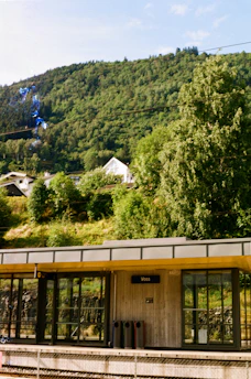 A small, open train station platform with a modern design is set against a lush, green hillside. In the background, multiple layers of thick forest stretch across the scene, while a few houses with slanted roofs are nestled among the trees. The clear blue sky enhances the peaceful atmosphere.