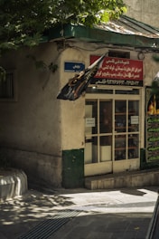 A small shop with a glass door and a red sign above it is situated at the corner of a building. There's a street sign reading 'Nayesh Alley' in blue. A black flag with an intricate design is suspended near the entrance. A tree with green leaves casts shadows onto the pavement and building, mostly shaded with some light spots visible.
