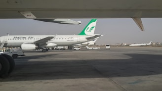 An airport apron with a parked Mahan Air airplane. The aircraft is a white Airbus A300 with a green logo on its tail. Various ground service vehicles, including a passenger bus, are nearby. Another airplane is seen taxiing on a runway in the background.