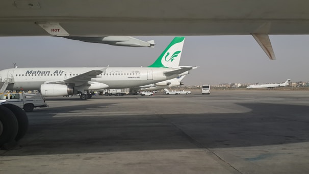 An airport apron with a parked Mahan Air airplane. The aircraft is a white Airbus A300 with a green logo on its tail. Various ground service vehicles, including a passenger bus, are nearby. Another airplane is seen taxiing on a runway in the background.