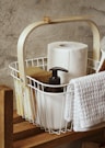 A wooden shelf holds a white wire basket filled with household items. Inside the basket, there is a roll of paper towels, a black pump soap dispenser, a sponge, and a white towel draped over the side. A dried flower is decoratively placed on the handle of the basket. The background features a textured grey concrete wall.
