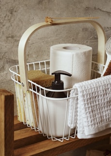 A wooden shelf holds a white wire basket filled with household items. Inside the basket, there is a roll of paper towels, a black pump soap dispenser, a sponge, and a white towel draped over the side. A dried flower is decoratively placed on the handle of the basket. The background features a textured grey concrete wall.
