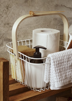 A wooden shelf holds a white wire basket filled with household items. Inside the basket, there is a roll of paper towels, a black pump soap dispenser, a sponge, and a white towel draped over the side. A dried flower is decoratively placed on the handle of the basket. The background features a textured grey concrete wall.
