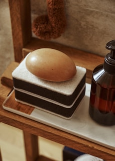 A brown oval-shaped soap rests on a black and white stacked sponge on a wooden shelf. Next to it, there is an amber-colored pump bottle. The setting is rustic and minimalistic, with warm tones and soft lighting.