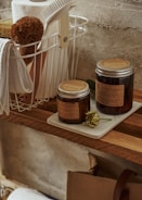 A wooden shelf holds two brown glass jars labeled 'Aroma Sensation' with silver lids, placed on a white tray alongside a dried green flower. Above the jars, a white metal basket contains a brown brush, a roll of paper towels, a sponge, a white cloth, and a white scrubbing brush with a wooden handle. The background features a textured concrete wall.