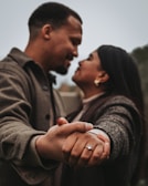A couple is intimately holding hands, focusing on the woman’s hand adorned with an engagement ring. The two individuals are looking into each other’s eyes, suggesting a moment of connection and affection.