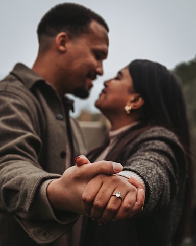 A couple is intimately holding hands, focusing on the woman’s hand adorned with an engagement ring. The two individuals are looking into each other’s eyes, suggesting a moment of connection and affection.