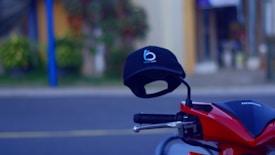 A black cap with a logo is placed on the handlebars of a red Honda motorcycle. The background shows a blurred street scene with foliage and buildings.