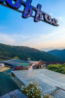 A scenic view featuring rolling hills covered with dense greenery under a clear blue sky. The foreground shows rooftops adorned with colorful flowers. A sign with the word 'coffee' is partially visible at the top, suggesting the presence of a café nearby. Mountains are faintly visible in the background, adding depth to the image.