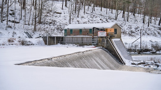 A snow-covered landscape with a small dam and control house situated in a forested area. The water flows over the dam while a danger sign is prominently displayed. Leafless trees line the hillside, and the sky is overcast, suggesting a cold winter day.