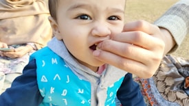A young child wearing a blue jacket with gray and white patterns is being fed by an adult's hand. The child appears to be sitting in an outdoor setting. Someone in the background is holding a phone and wearing a patterned attire.