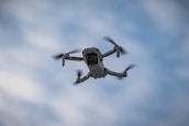 A gray drone with four propellers is flying in the sky. The sky is overcast with a light blue and grayish tone, indicating it might be a cloudy day.