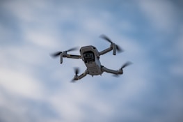 A gray drone with four propellers is flying in the sky. The sky is overcast with a light blue and grayish tone, indicating it might be a cloudy day.