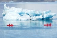Two red kayaks with people wearing red gear paddle in a calm, icy body of water, passing by a large, white and blue iceberg under an overcast sky.