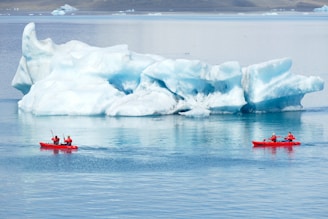 Two red kayaks with people wearing red gear paddle in a calm, icy body of water, passing by a large, white and blue iceberg under an overcast sky.