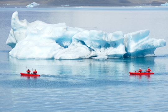 Two red kayaks with people wearing red gear paddle in a calm, icy body of water, passing by a large, white and blue iceberg under an overcast sky.