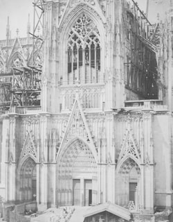 A detailed gothic cathedral facade featuring intricate carvings and statues, with pointed arches and large, ornate windows. Scaffolding suggests ongoing construction or restoration work.