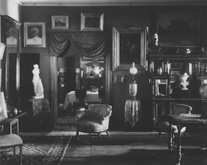 An ornate, vintage interior filled with various framed paintings and sculptures. The room features plush chairs, a table draped with a cloth, and a standing lamp with intricate designs. Rich textures and patterns are present in the carpets and curtains, adding to the classical ambiance. A statue is prominently displayed next to a doorway leading to a dining area.