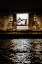A large illuminated advertisement is displayed on the facade of an ornate building at night. The advertisement features a person lounging on a chair with luggage nearby, promoting a luxury brand. The building has arched windows and intricate stone carvings, illuminated by spotlights. Reflections of city lights glisten on the rippling water in the foreground.