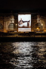 A large illuminated advertisement is displayed on the facade of an ornate building at night. The advertisement features a person lounging on a chair with luggage nearby, promoting a luxury brand. The building has arched windows and intricate stone carvings, illuminated by spotlights. Reflections of city lights glisten on the rippling water in the foreground.