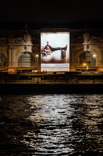 A large illuminated advertisement is displayed on the facade of an ornate building at night. The advertisement features a person lounging on a chair with luggage nearby, promoting a luxury brand. The building has arched windows and intricate stone carvings, illuminated by spotlights. Reflections of city lights glisten on the rippling water in the foreground.
