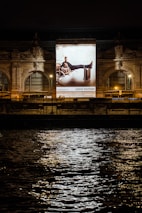 A large illuminated advertisement is displayed on the facade of an ornate building at night. The advertisement features a person lounging on a chair with luggage nearby, promoting a luxury brand. The building has arched windows and intricate stone carvings, illuminated by spotlights. Reflections of city lights glisten on the rippling water in the foreground.
