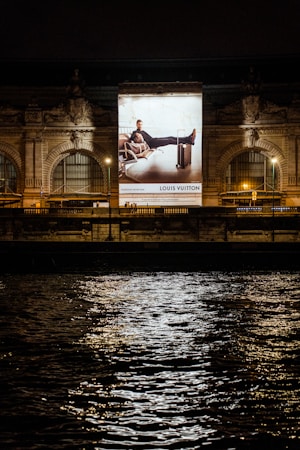 A large illuminated advertisement is displayed on the facade of an ornate building at night. The advertisement features a person lounging on a chair with luggage nearby, promoting a luxury brand. The building has arched windows and intricate stone carvings, illuminated by spotlights. Reflections of city lights glisten on the rippling water in the foreground.