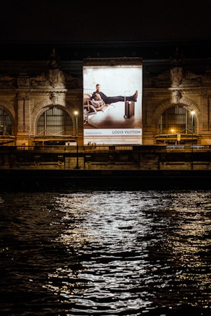 A large illuminated advertisement is displayed on the facade of an ornate building at night. The advertisement features a person lounging on a chair with luggage nearby, promoting a luxury brand. The building has arched windows and intricate stone carvings, illuminated by spotlights. Reflections of city lights glisten on the rippling water in the foreground.