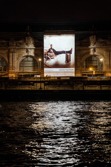 A large illuminated advertisement is displayed on the facade of an ornate building at night. The advertisement features a person lounging on a chair with luggage nearby, promoting a luxury brand. The building has arched windows and intricate stone carvings, illuminated by spotlights. Reflections of city lights glisten on the rippling water in the foreground.