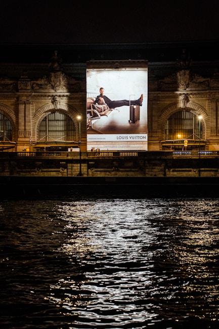 A large illuminated advertisement is displayed on the facade of an ornate building at night. The advertisement features a person lounging on a chair with luggage nearby, promoting a luxury brand. The building has arched windows and intricate stone carvings, illuminated by spotlights. Reflections of city lights glisten on the rippling water in the foreground.