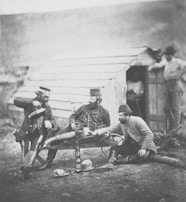 Several men in military attire are gathered around a table outside a rustic wooden structure. One man is pouring a drink while others are seated, casually interacting with each other. Additional figures are visible, leaning against the building, observing the scene. Various items, including bottles and hats, are scattered on the ground.