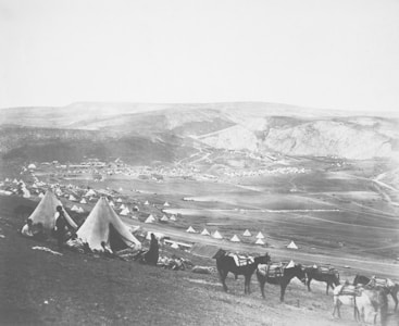 A vast landscape featuring numerous tents scattered across rolling hills, with a few people visible near the tents in the foreground. Several pack animals, possibly donkeys, are positioned nearby. The terrain stretches toward distant hills under a clear sky.