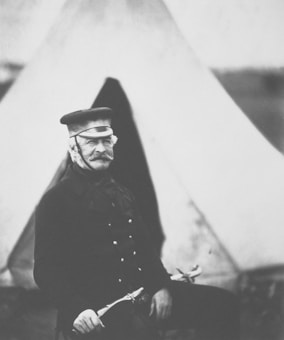 A black and white portrait of an older man with a mustache, wearing a uniform with buttons and a hat, seated in front of a tent. He holds a sword in one hand and gazes directly at the camera, conveying a sense of composure and authority.