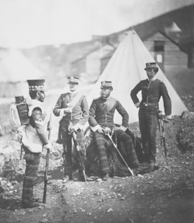 Four men dressed in military uniforms stand and sit together outdoors. Three of them are standing, and one is seated on a piece of unidentified material. The men are wearing ornate hats and what appear to be kilts or tartan pants. In the background, there are tents and wooden structures with a hilly landscape visible.