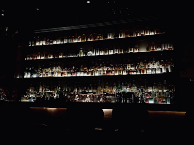 A moody, dimly lit bar shelf showcasing an array of aged genever bottles with deep green and gold accents.