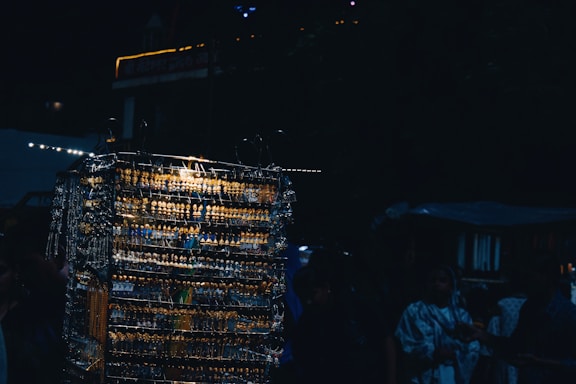 A dimly lit outdoor market scene featuring a display of multiple earrings and necklaces arranged on racks. The metal jewelry reflects light, creating a glimmering effect. Several people, partially obscured by shadows, are browsing the items.