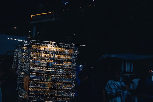 A dimly lit outdoor market scene featuring a display of multiple earrings and necklaces arranged on racks. The metal jewelry reflects light, creating a glimmering effect. Several people, partially obscured by shadows, are browsing the items.