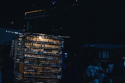 A dimly lit outdoor market scene featuring a display of multiple earrings and necklaces arranged on racks. The metal jewelry reflects light, creating a glimmering effect. Several people, partially obscured by shadows, are browsing the items.