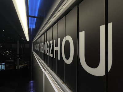 A close-up view of a large, black and white sign displaying letters spelling out 'ZHENGZHOU', illuminated by overhead fluorescent lighting. The background reveals a dimly lit interior, possibly indicating an airport or train station setting.