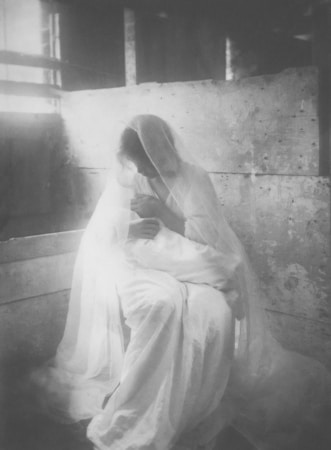 A woman dressed in a flowing white garment, possibly a veil, is sitting in a dimly lit room with rustic wooden panels. The scene exudes a sense of calm and serenity, with soft lighting creating a gentle ambiance.