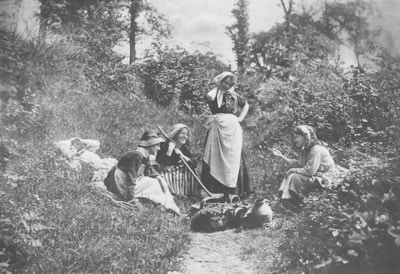 Four women are gathered in a rural setting, dressed in traditional clothing with bonnets and aprons. They are seated or standing on a grassy path surrounded by lush vegetation. The scene includes several clay pots and baskets filled with produce.